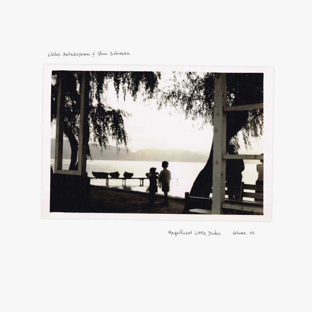 Washed out black and white photo of two young children standing at the edge of a large lake with mountains behind them, hazy from the setting sun. There are old trees around them and some other partially obstructed people near an old white deck type structure.