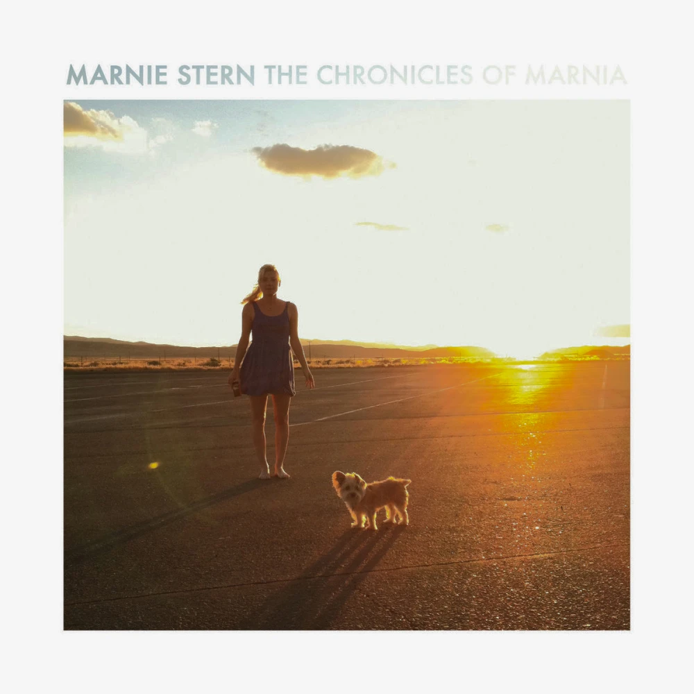 Photograph of Marnie, a white barefoot woman in a short dress with long blonde hair in a pony tail. She is standing on blacktop next to a small breed dog with a desert landscape behind her. The sun is setting, causing a blinding effect overall. Her name and the record title are set above the photo in a light font that fades to transparent from left-to-right.