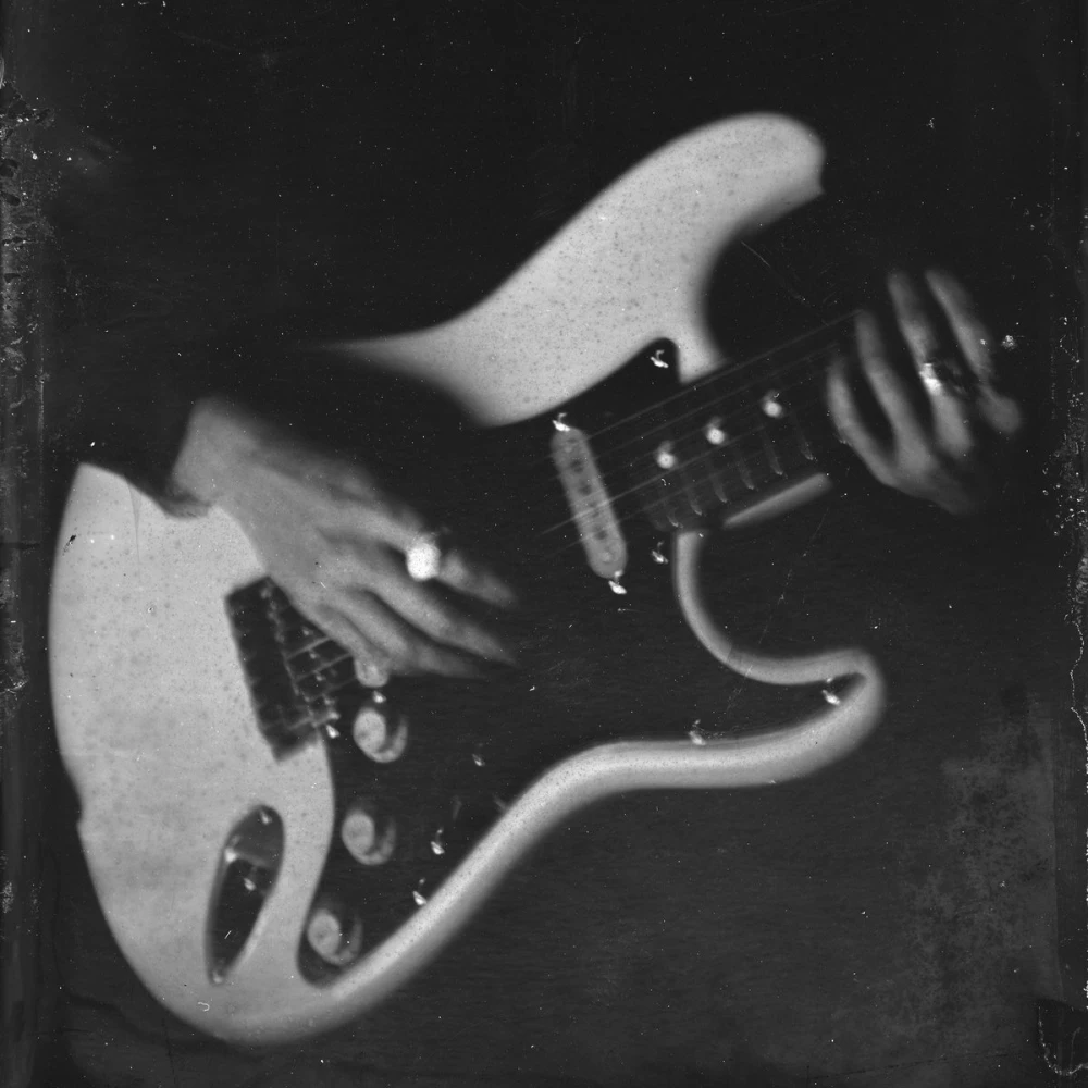 Grainy black and white photo of a woman’s hands with rings on her fingers, playing an electric guitar.