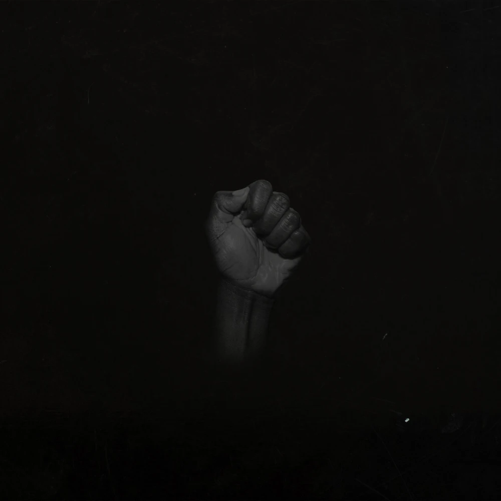 Very dim raised fist with brown skin in a resistance and protest pose, over a solid black background.