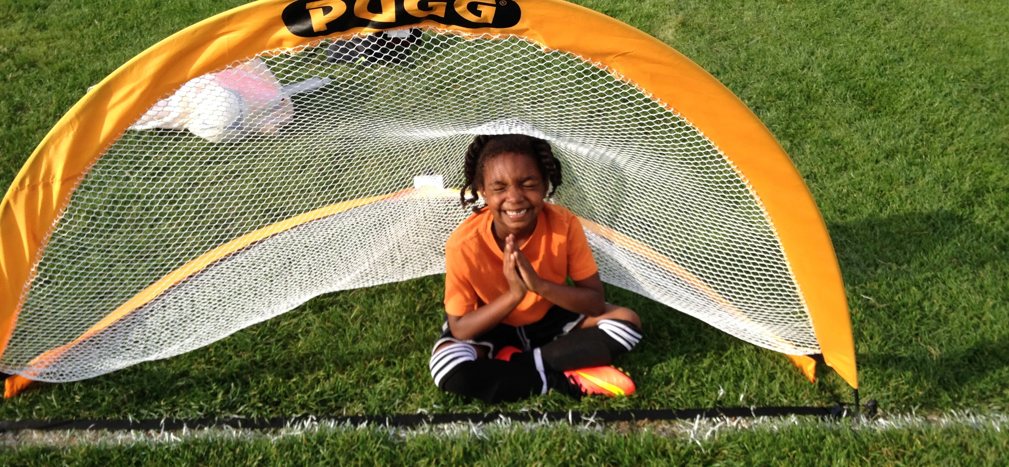 Young Black girl with braided hair, wearing a soccer kit and sitting cross legged in a Pugg soccer goal with her hands folded in a meditation pose while grinning widely.