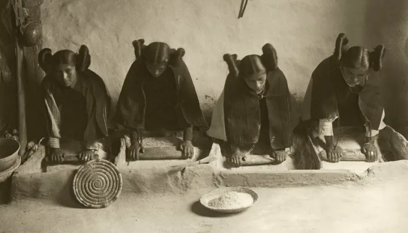 Four Hopi women in a row, kneeling on dirt floor in a house, rolling pins over grain. They are wearing traditional clothing and their shared hairstyle is center parted with very large extended buns on the sides of their heads.