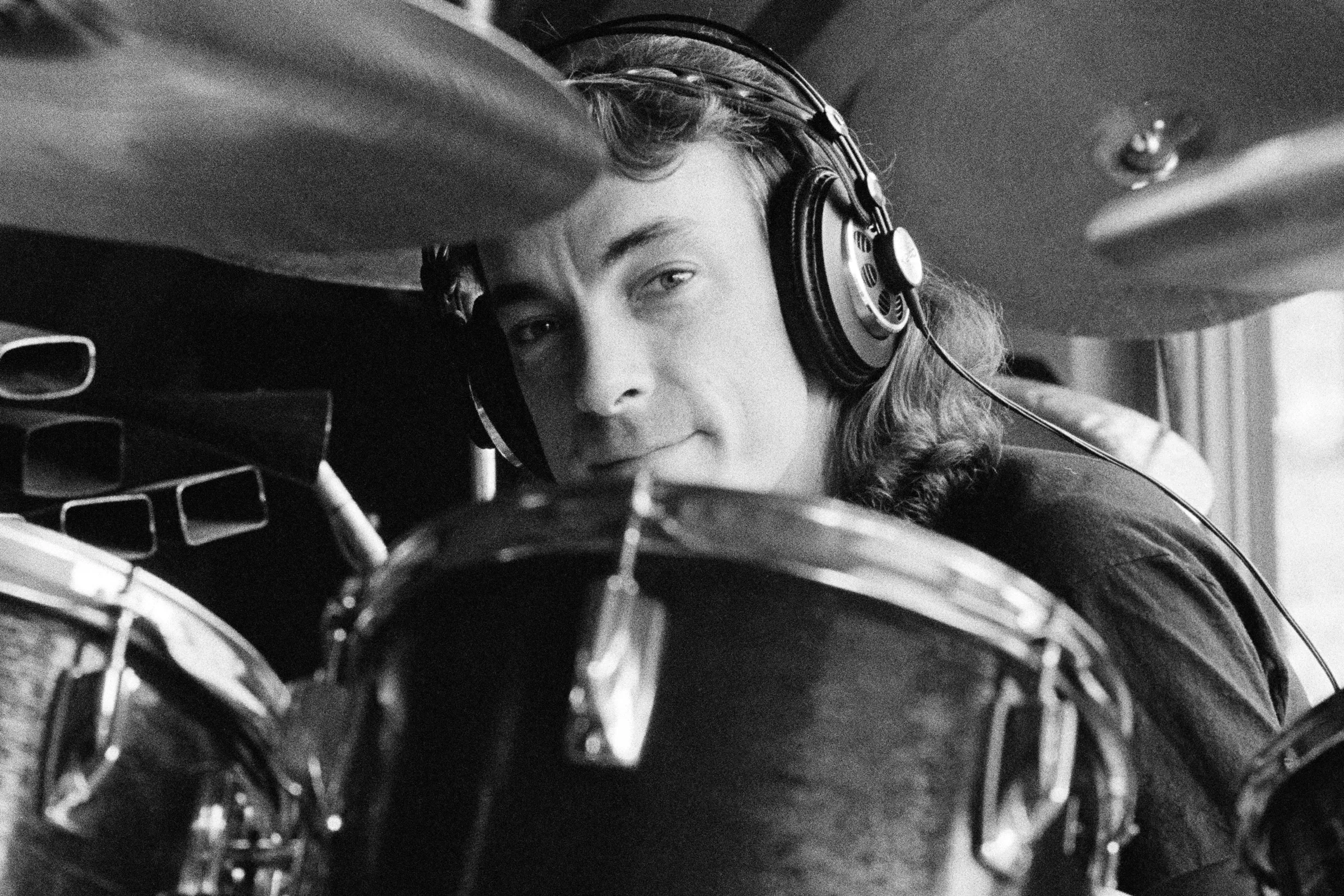 A young Neil with shoulder length hair, wearing headphones, peaking out from behind the small spaces between his drums and cymbals, with a pleasant smile.