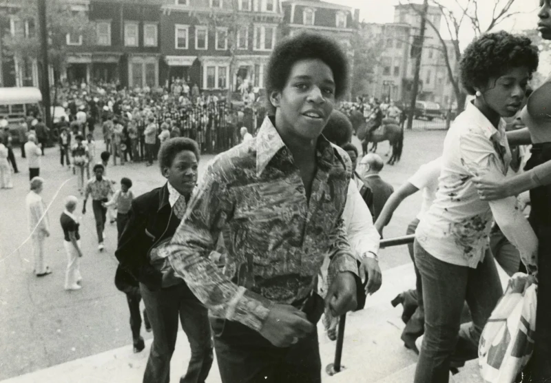 Scene in front of a school with a group of Black students climbing the steps as they enter the building. Front and center is a boy with a large afro and a vividly-patterned dress shirt with a butterfly collar. Near him is a girl in a nice flowery shirt with a concerned look on her face, being comforted by an adult. Behind him some other students are approaching, and behind them are a sea of white people near an iron fence, protesting their approach.