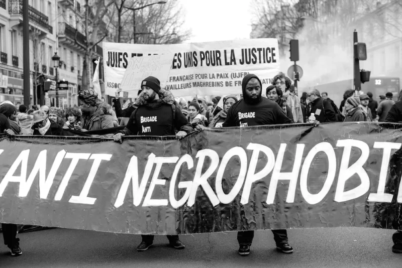 Protestors in the street. The leaders are wearing black shirts and carrying a large banner, all donning the label 'brigade anti-négrophobie.’