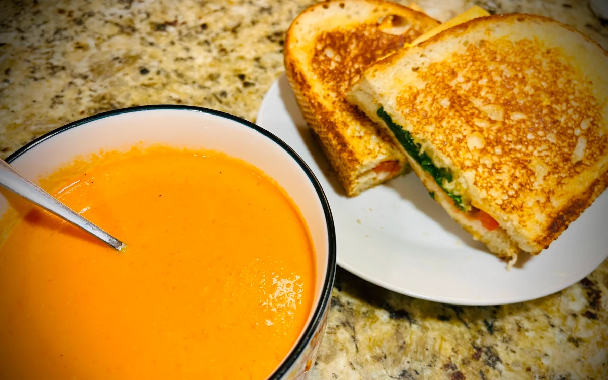 Bright orange creamy smooth soup in a bowl next to a grilled sandwich cut in two on a plate. The green spinach, red tomato, and meaty vegan cheese are protruding from the sliced side.