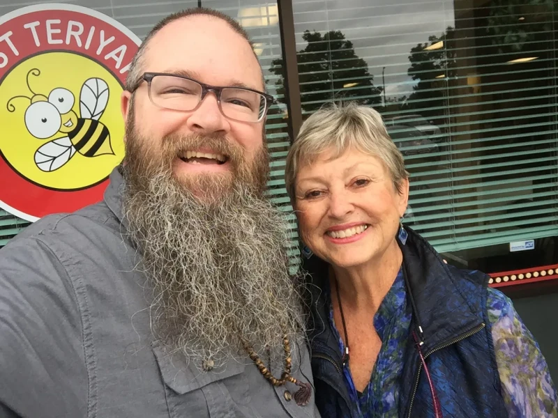 Mrs. Smith and me, smiling in a selfie outside in front of a restaurant window. I have glasses and a long beard, and I'm wearing a dress shirt and mala beads. Mrs. Smith has short hair and is wearing a vest jacket over a long-sleeve shirt.