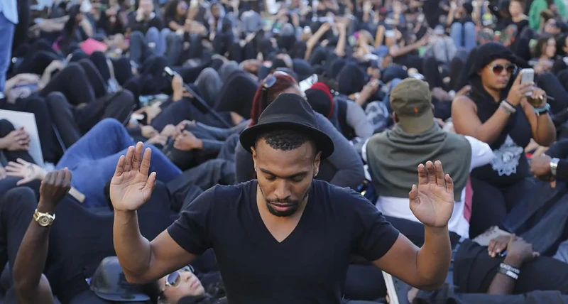 Scene of a protest with a crowd of people (mostly Black) lying down on the ground. In the front, one Black man in a t-shirt and fedora is kneeling and looking at the ground with his hands raised as if being confronted by police.
